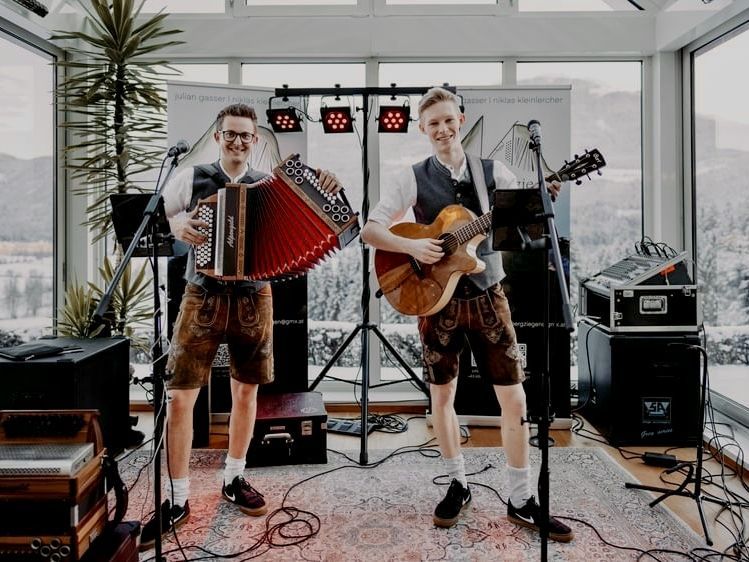 Zwei junge Musiker in traditioneller Tracht spielen in einem lichtdurchfluteten Raum mit Alpenpanorama im Hintergrund. Einer spielt Akkordeon, der andere Gitarre, beide stehen l&auml;chelnd auf einem Teppich vor Mikrofonen und Lautsprechern.
