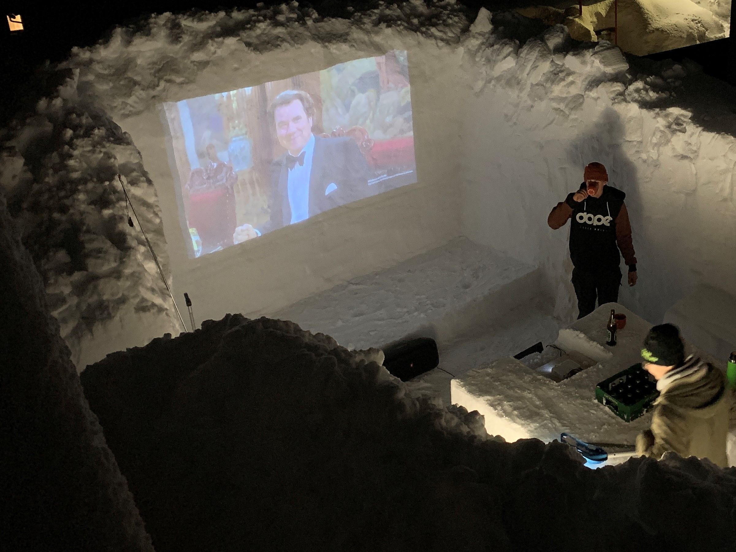 Eine Schneewand dient als Leinwand, in der Mitte steht ein selbstgemachter Tisch f&uuml;r Getr&auml;nke.