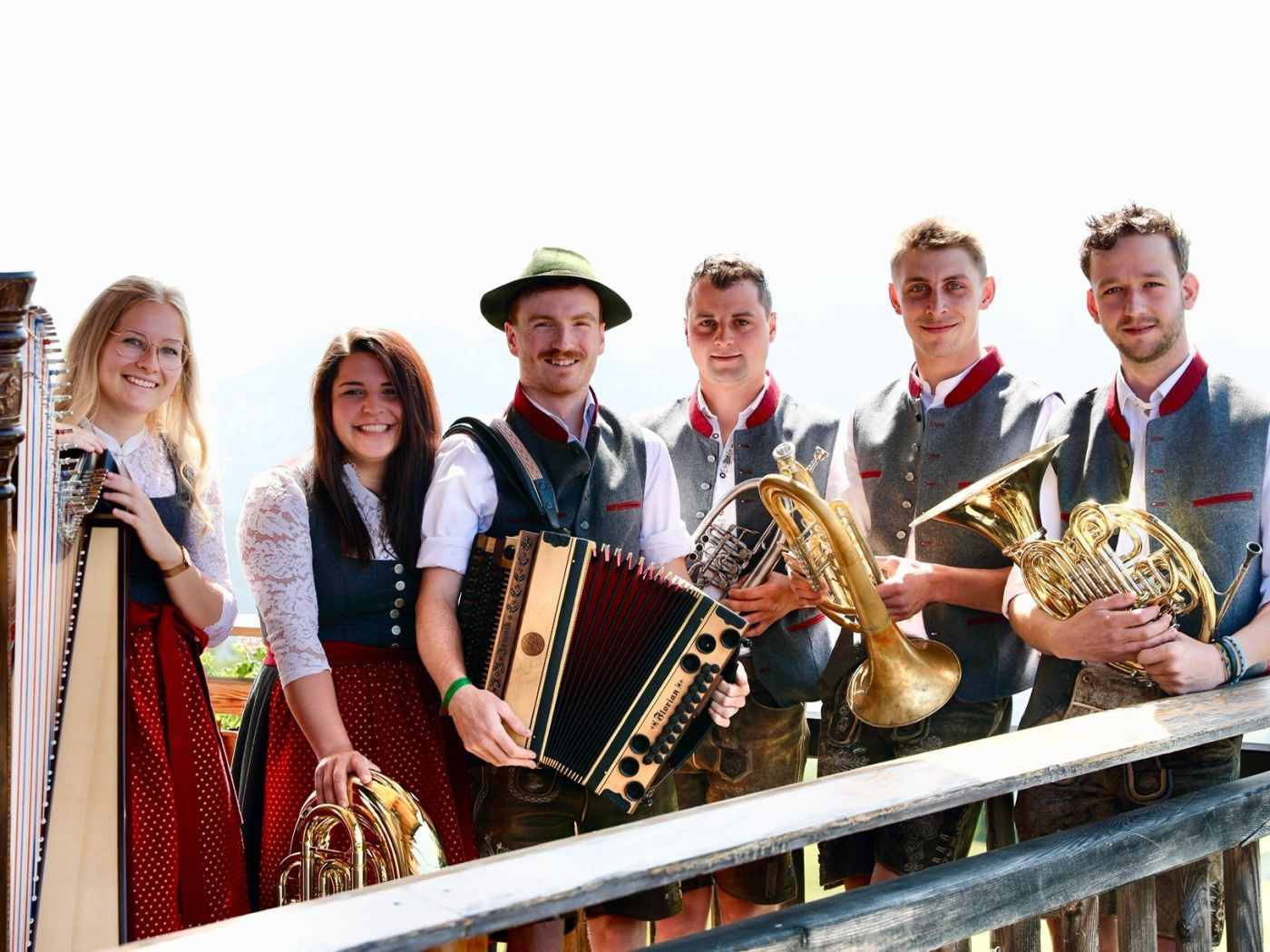 Sechs Musiker in traditioneller Tracht mit Instrumenten wie Harfe, Akkordeon, Trompete und Hörnern, lächelnd im Freien vor Bergkulisse